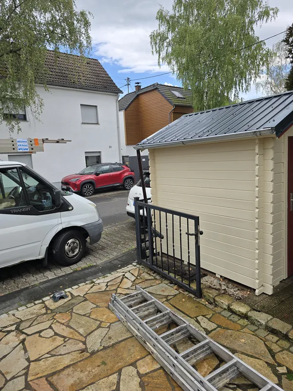Gartenhaus mit neuem dunklem Blechdach, davor eine Leiter auf dem Plattenweg