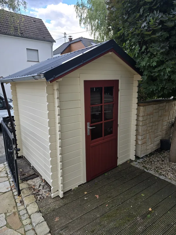 Fertiges Gartenhaus mit hellem Anstrich, roter Tür und dunklem Dach